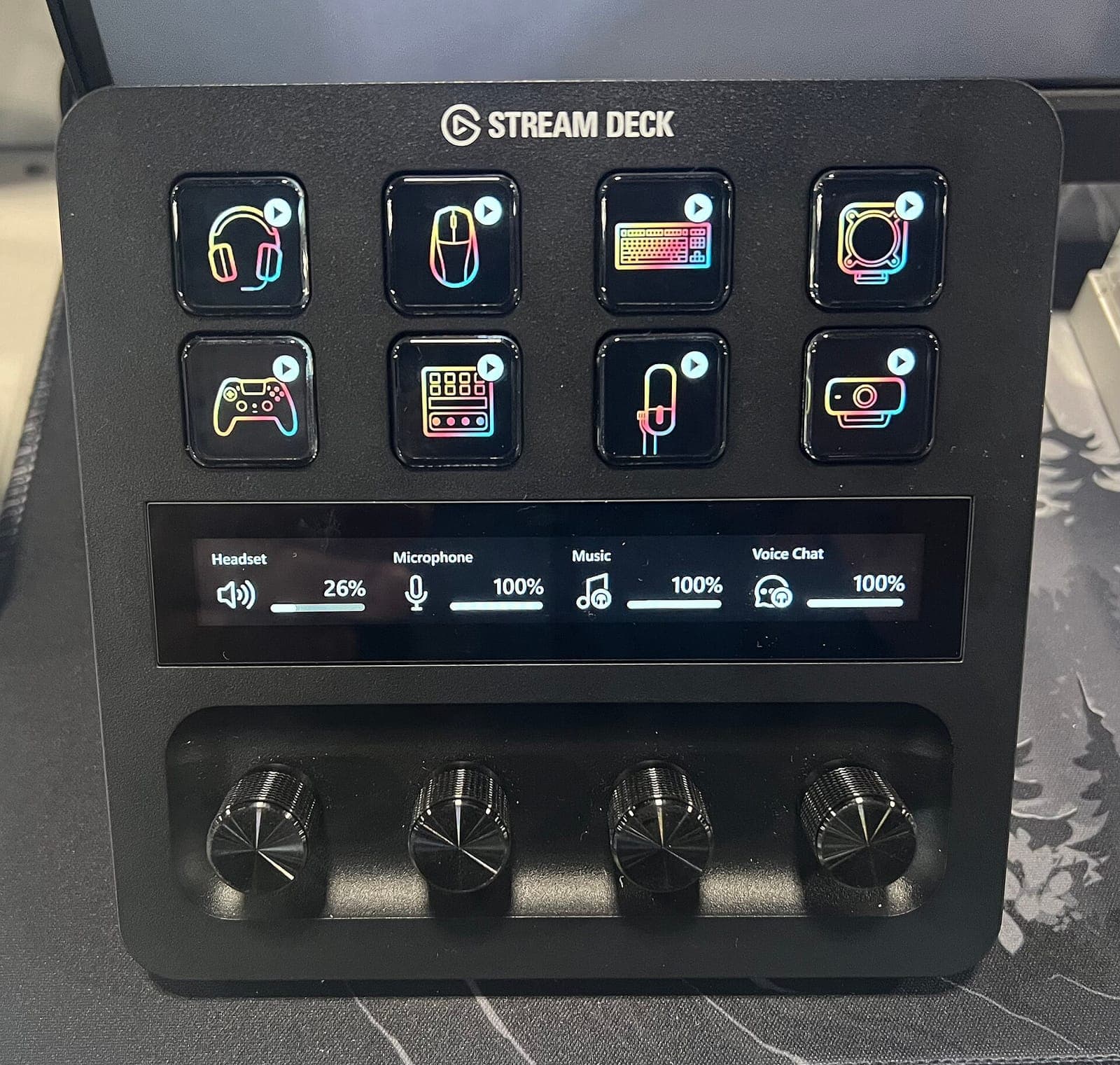 An Elgato Stream Deck + on a desk with eight lit keys, a touch strip showing audio levels and four dials.